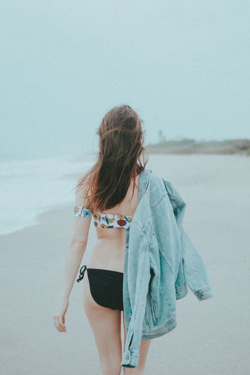 Free Woman strolling on a beach in a bikini with a denim jacket, misty backdrop. Stock Photo