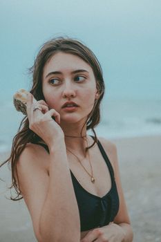 A young woman in a black bikini holds a seashell by the ocean on a cloudy day.