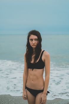 A young woman stands on a beach wearing a black bikini, under a cloudy sky.