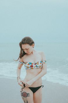A woman with a leg tattoo stands on the beach in a colorful bikini.