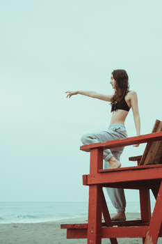 Teen girl in casual wear points from a red lifeguard stand on a cloudy beach.