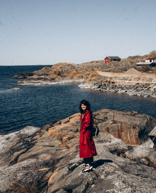 Free A young woman in a red coat standing on rocky coast by the sea on a sunny day. Stock Photo