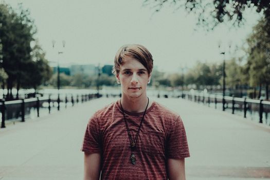 A young man in a red shirt with necklaces stands in a city walkway, showcasing a casual urban style.