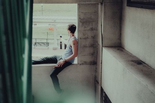 Free Young man in casual outfit sits on a windowsill, enjoying a city view, capturing urban calmness. Stock Photo