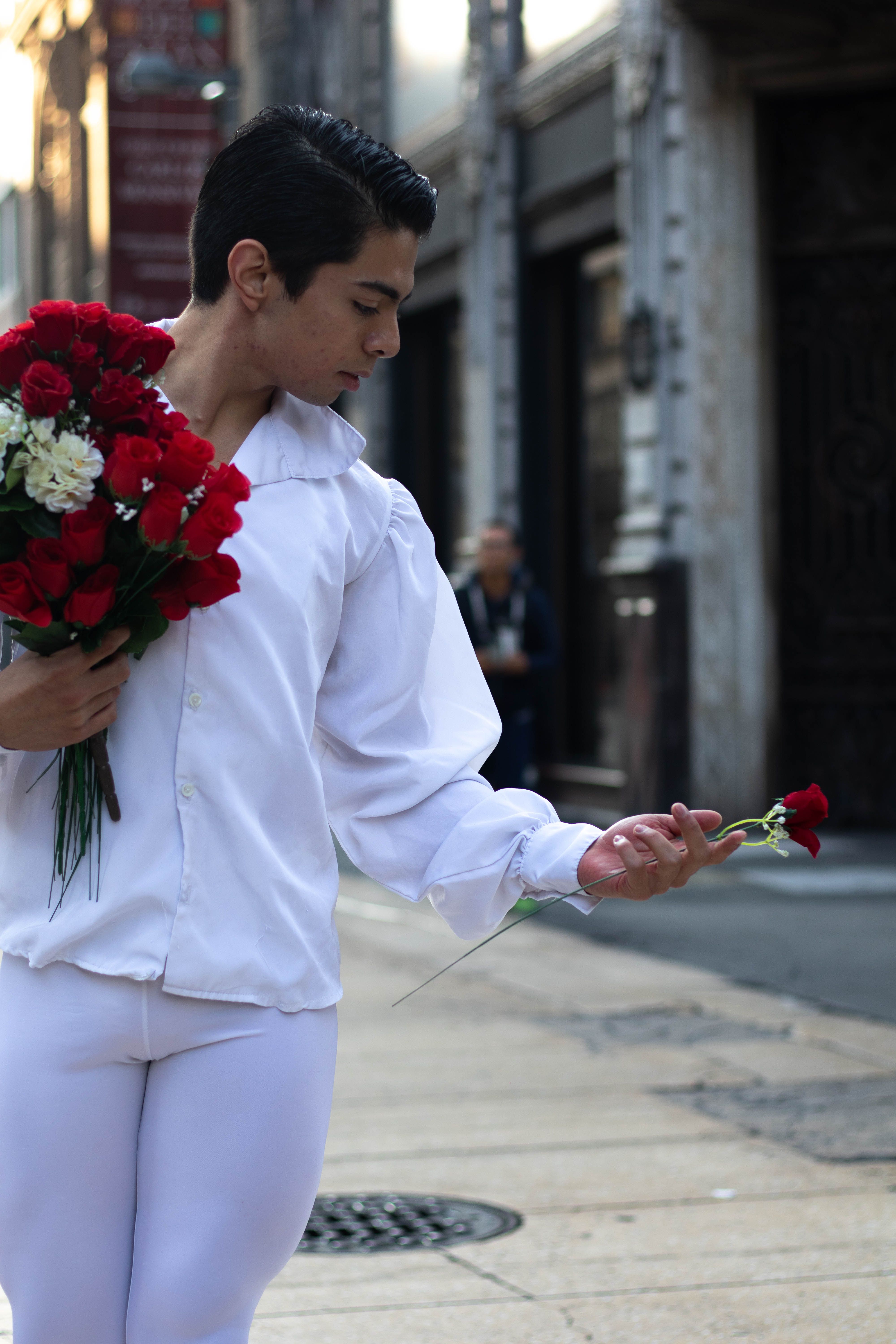A young man in a white outfit holds a bouquet of red roses in a street setting, showcasing elegance.
