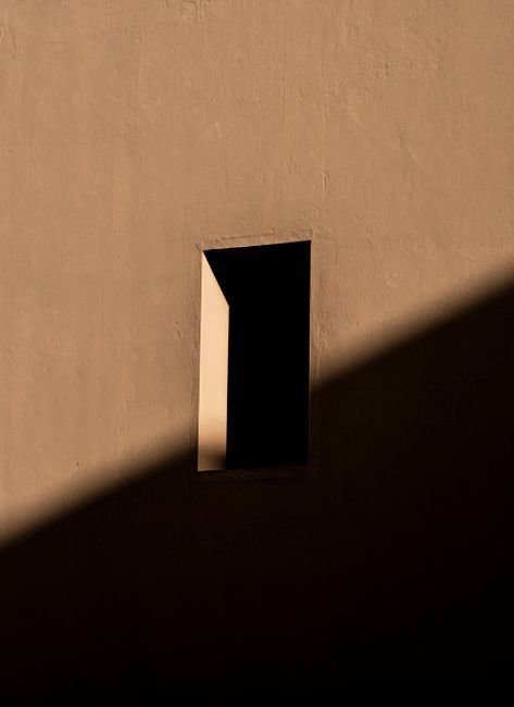 A minimalist view of a shadow creeping on a building wall with a small window, in warm light.