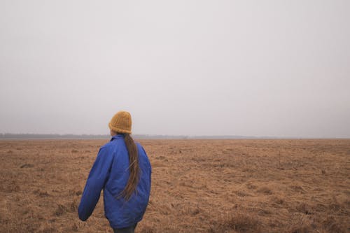 Person in Blue Jacket and Brown Knit Cap Walking on Brown Field