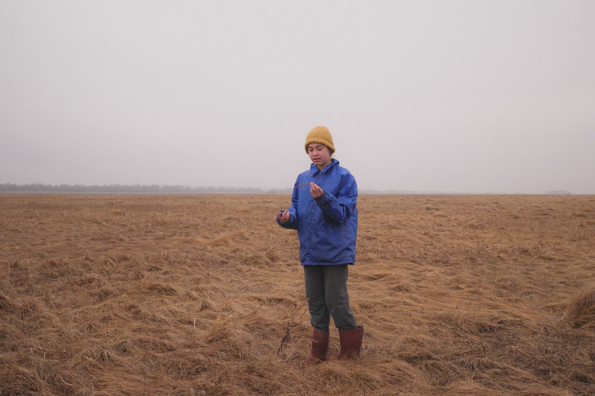 A young woman in a blue jacket and beanie stands in a vast dry field.