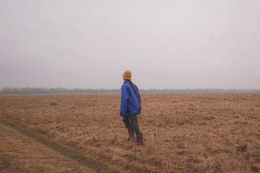 A young woman in a blue jacket and beanie stands in a foggy rural field, looking into the distance.
