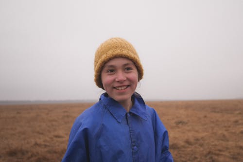 Smiling Young Woman in a Yellow Knitted Hat and Blue Raincoat Standing in the Field