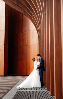 Bride and groom in elegant attire embracing by modern wooden architecture on their wedding day.