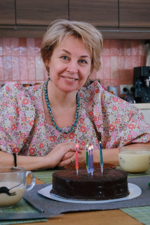 An Elderly Woman Wearing Floral Blouse Sitting Near the Cake with Lighted Candles while Smiling at the Camera