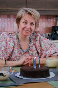 Elderly woman smiling near a chocolate cake with candles in a cozy kitchen.