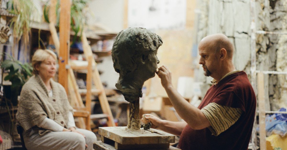 A sculptor shaping a clay head bust in an art studio with a woman observing.