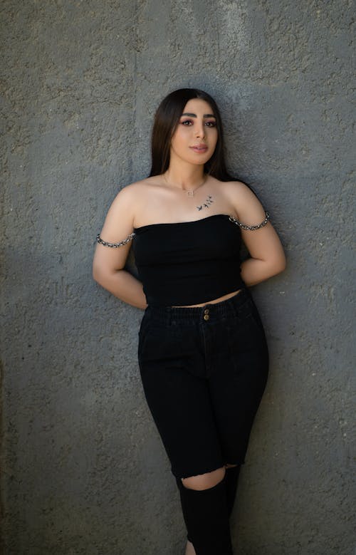 Free Woman in Black Tube Top and Black Pants Leaning on Gray Wall while Smiling at the Camera Stock Photo