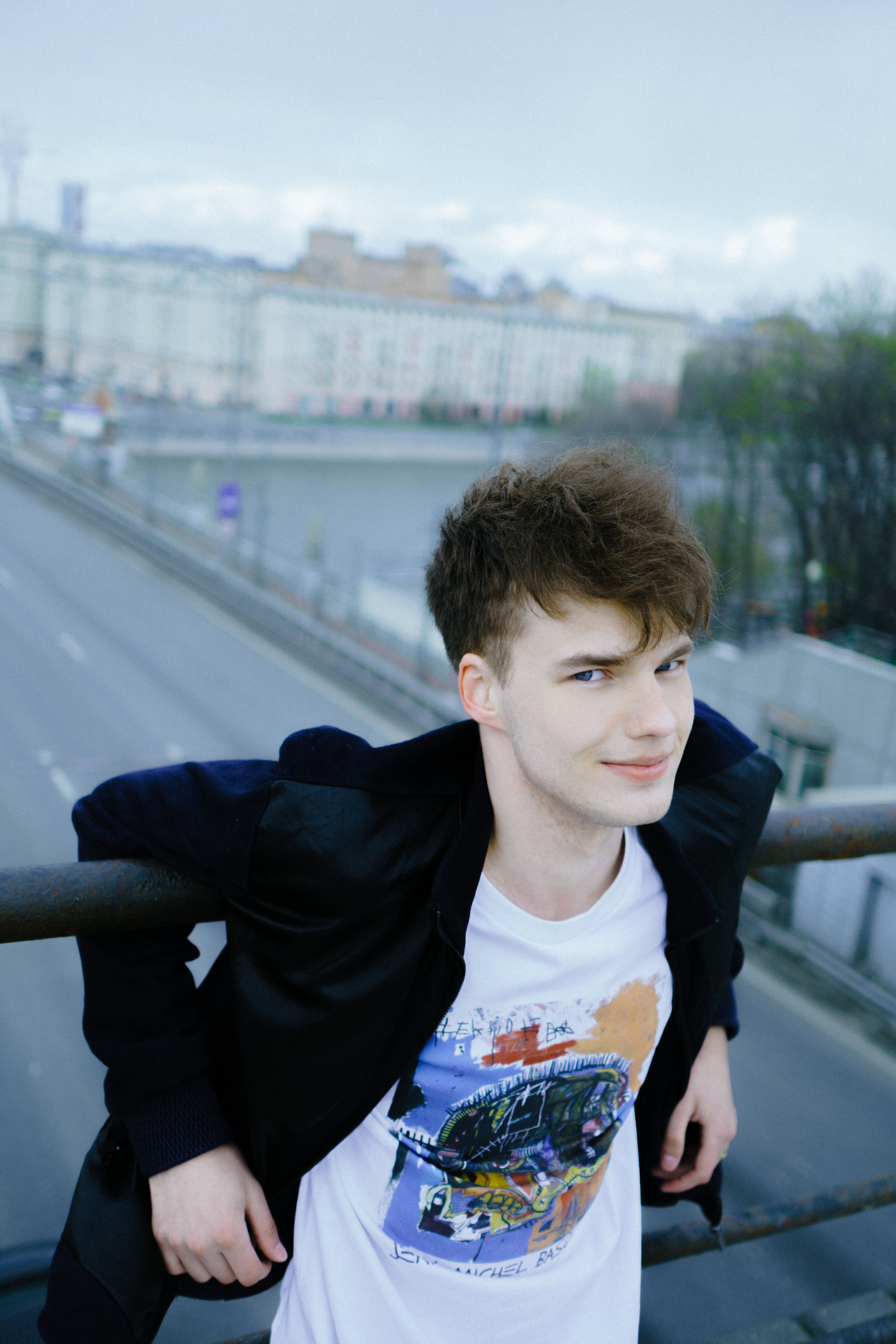 A young man in a black jacket leans on a railing overlooking a city.