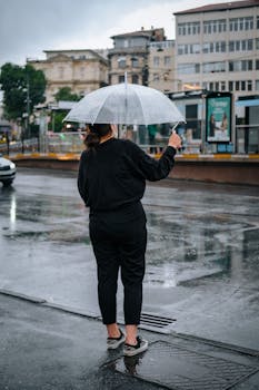A woman in a black outfit stands with a clear umbrella on a rainy sidewalk in İstanbul, Türkiye.