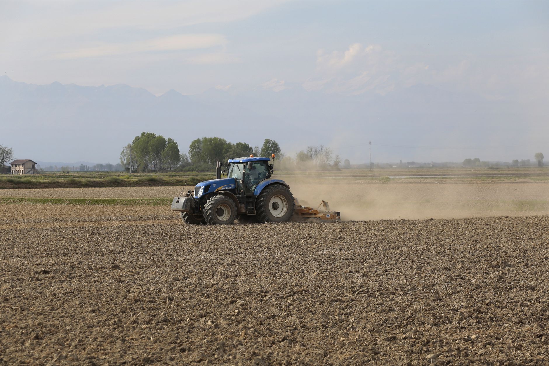 A blue tractor tills an expansive field on a sunny day, creating dust clouds.