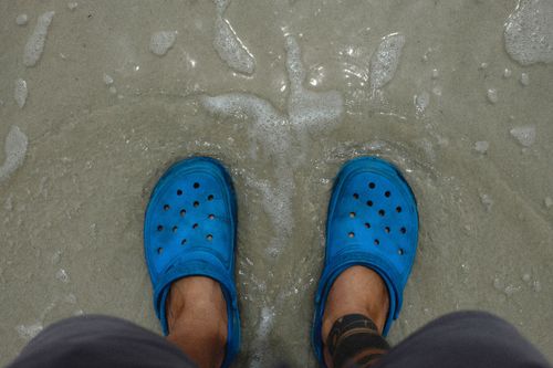 Free Top view of blue clogs on sandy beach with gentle waves lapping at feet. Stock Photo