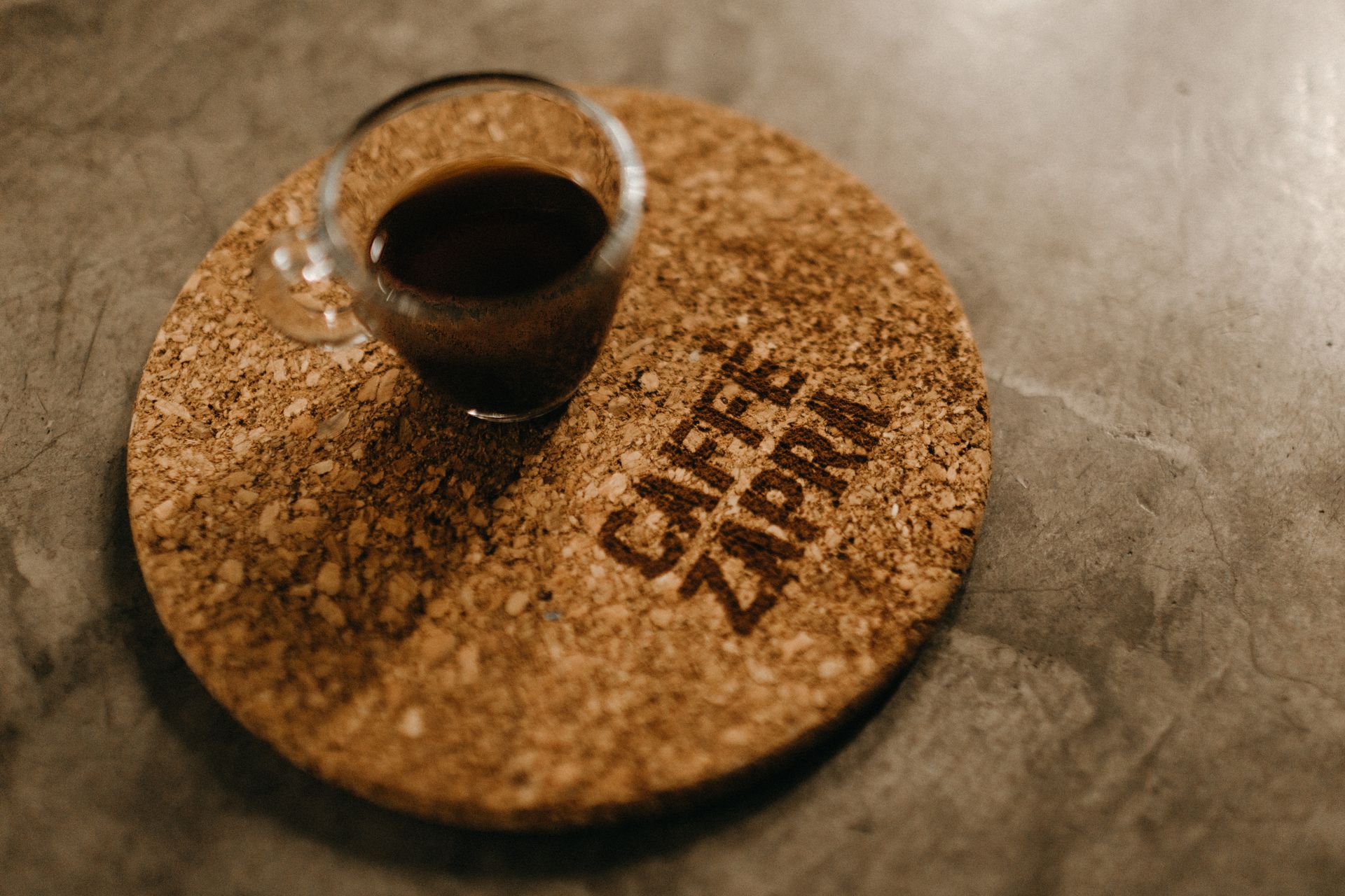 A cork coaster holding a glass of black coffee on a concrete surface.