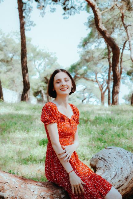 A young woman enjoys a sunny day in the park, sitting on a tree trunk with eyes closed and smiling.