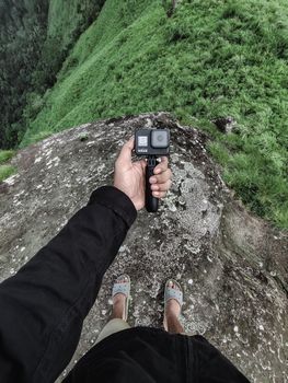 Man holding action camera over lush green cliff, ready to explore.