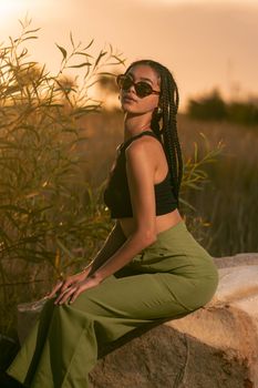 A fashionable woman with braided hair poses outdoors in a crop top and green pants during a stunning sunset.