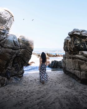 A woman in a long floral dress walks between big rocks on a sandy Mexican beach under a blue sky.