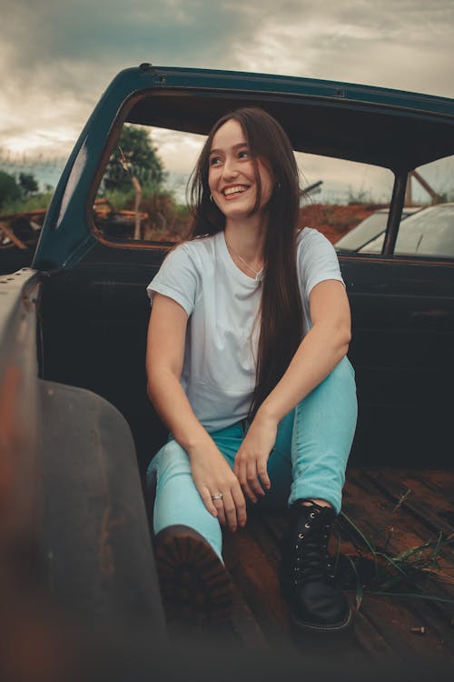 Free Woman in White Crew Neck T-shirt and Blue Denim Jeans Sitting on Black Car Stock Photo