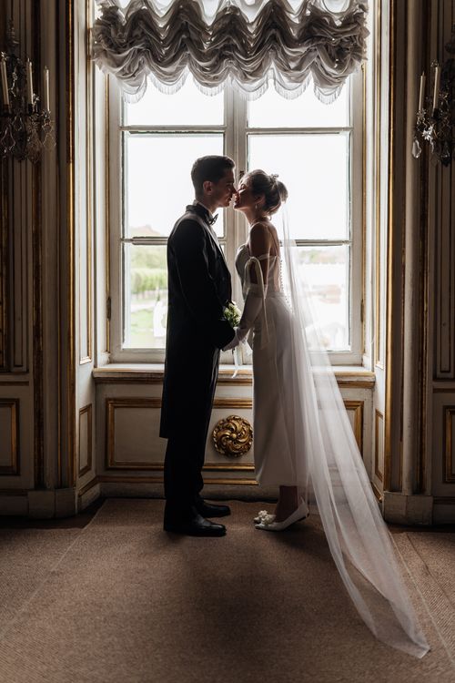 Free A bride and groom share a romantic kiss in an elegant interior setting, framed by a large window. Stock Photo