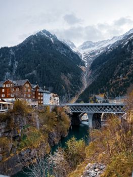 Picturesque view of Göschenen village with a bridge, river, and snow-capped mountains in autumn.