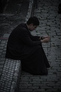 A priest in a cassock sitting on a cobblestone street, holding a rosary.