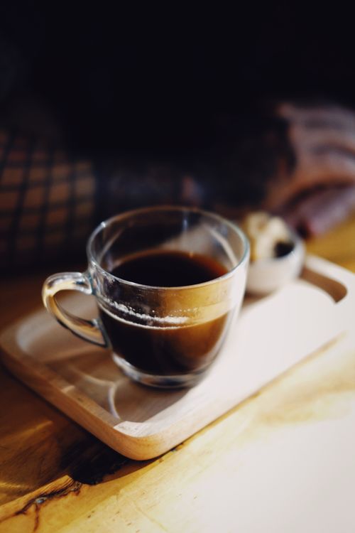 Free Close-up of a glass coffee cup on a wooden tray with a blurred tattooed arm in the background. Stock Photo