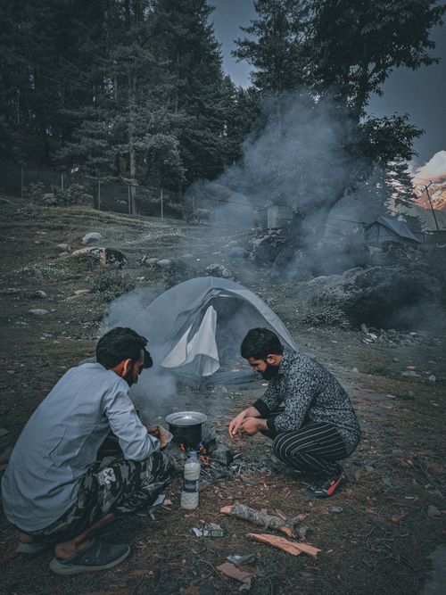 Free Two men cooking at a campsite with a tent set up in a forested area. Stock Photo