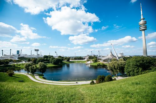 Free Beautiful day view of Munich's Olympic Tower reflecting in a picturesque lake surrounded by green parkland. Stock Photo