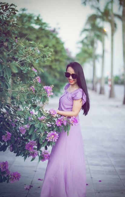 Elegant woman in a purple dress and sunglasses smiling outdoors by blooming flowers.