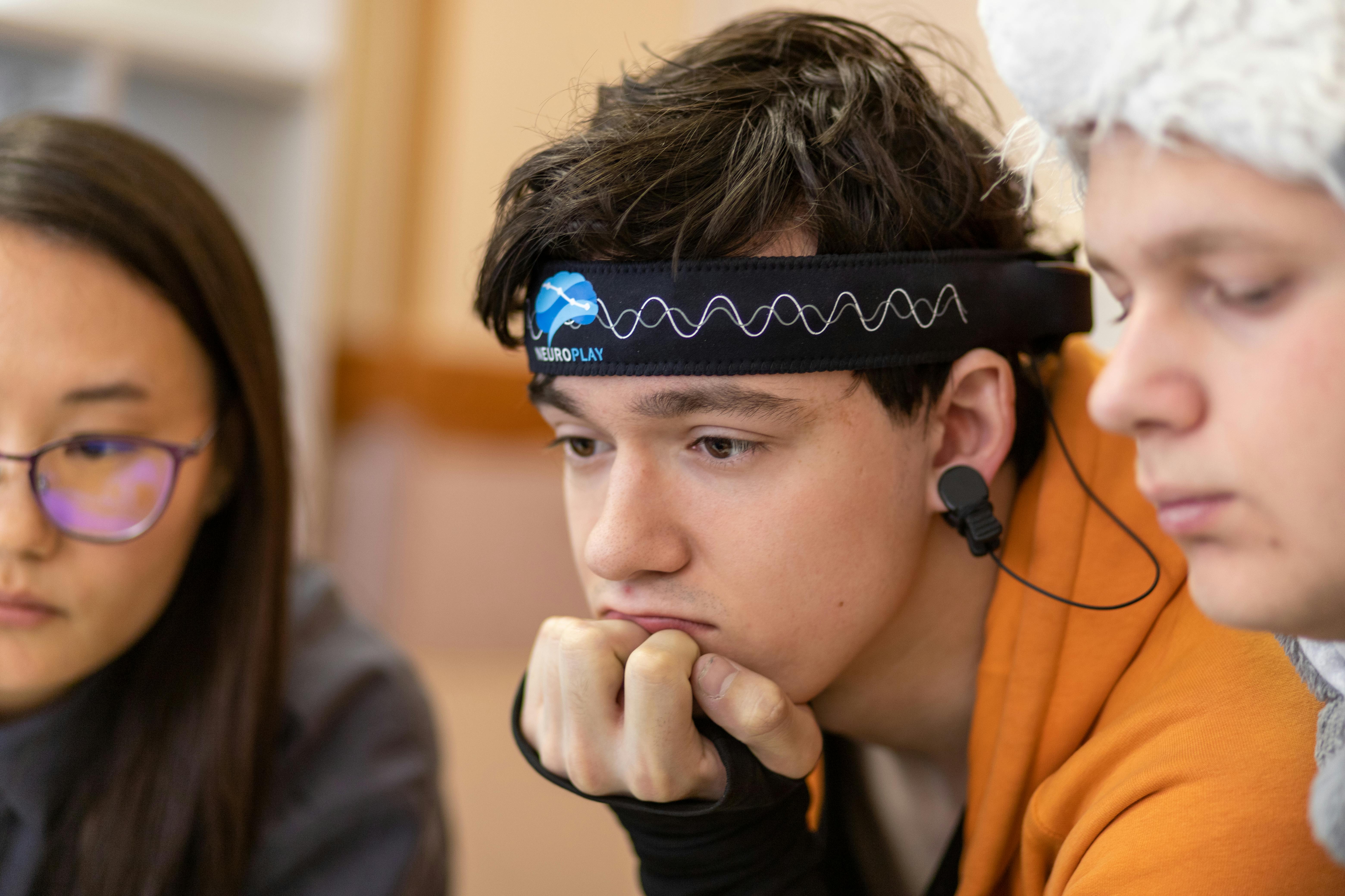 A young person with a headband device looks intently, resting chin on hand, alongside two others.