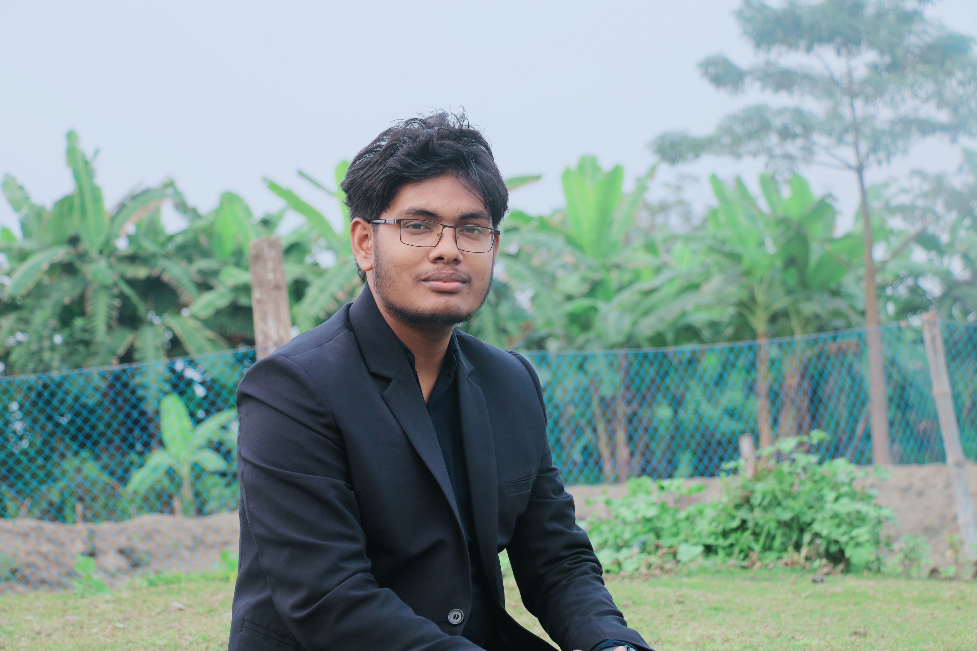 Confident young man in a black suit outdoors against a lush nature background.