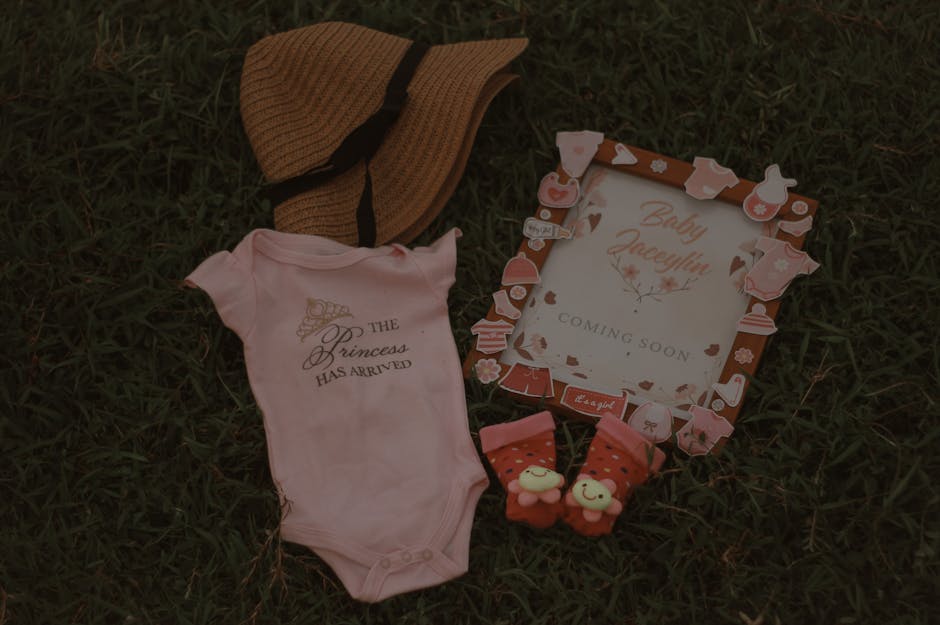 Flat lay of baby arrival announcement with pink onesie and accessories on grass.