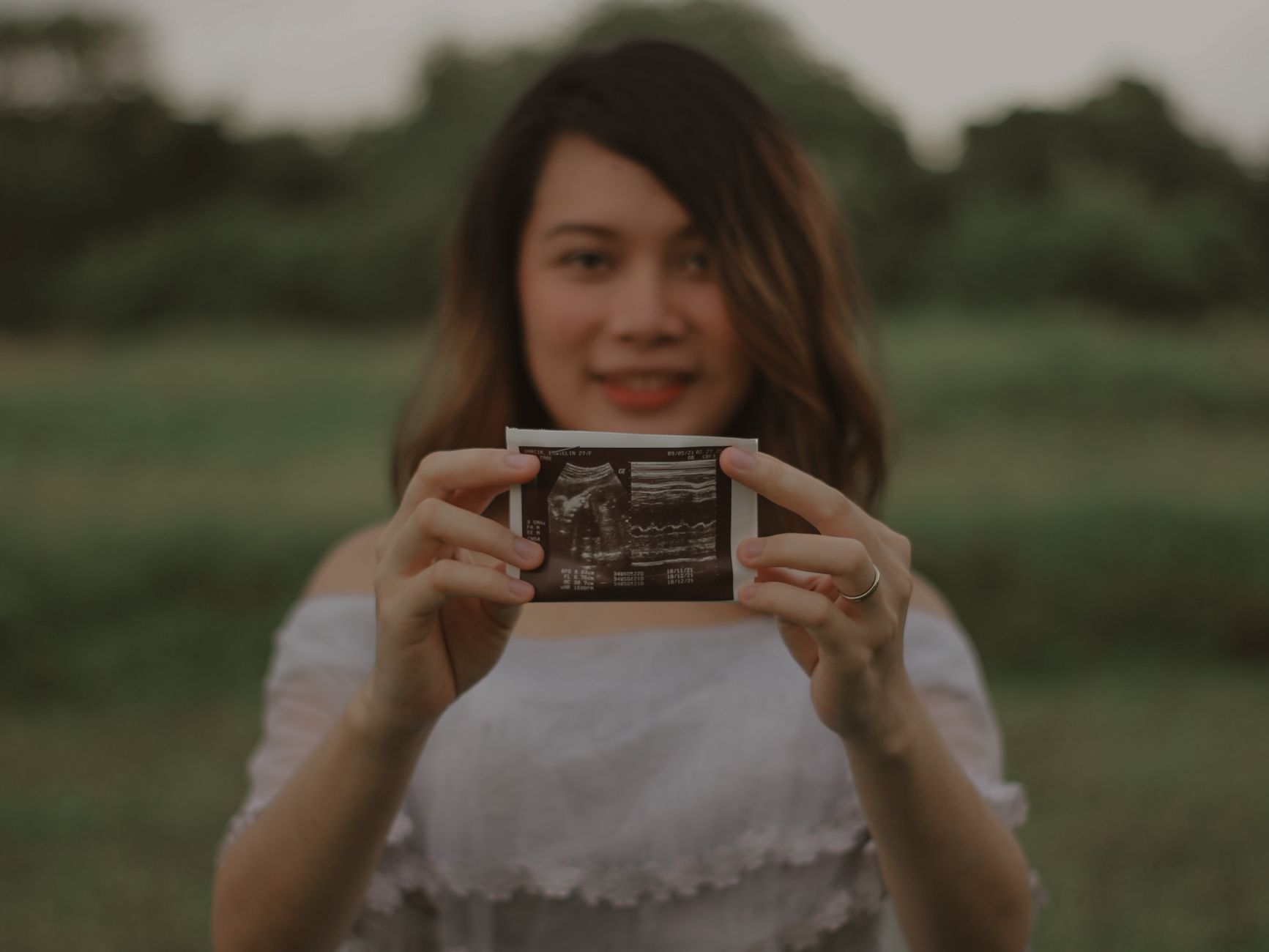 A smiling pregnant woman holds an ultrasound photo outdoors, symbolizing joy and anticipation.