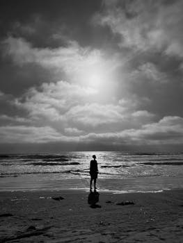 A person stands on the beach, silhouetted against a dramatic sea and sky in black and white.