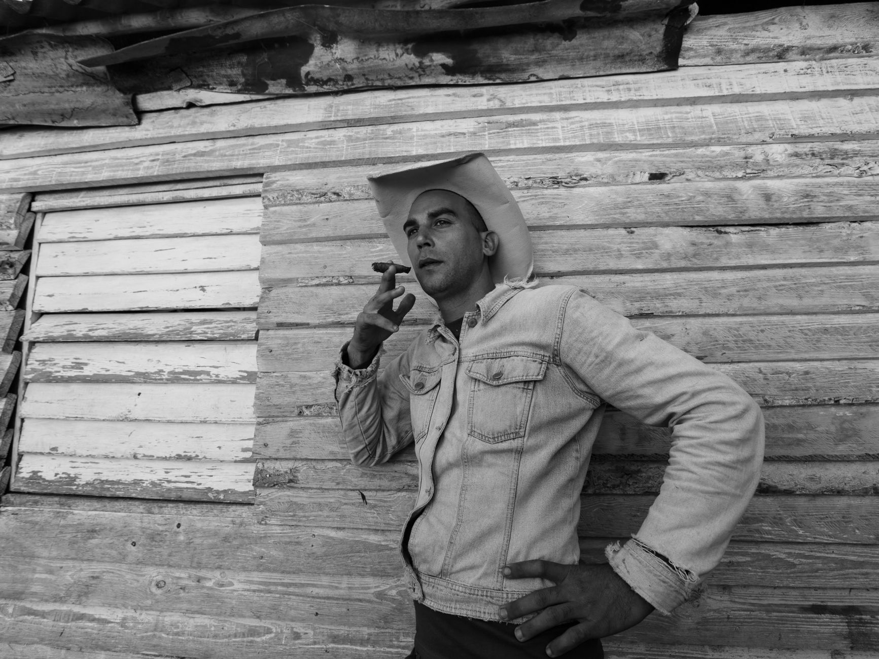 Black and white portrait of a cowboy in a hat, smoking outside a rustic wooden structure.
