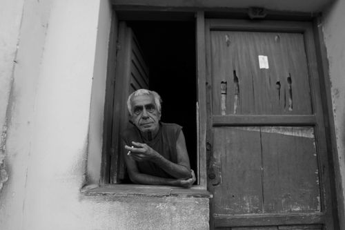 Free Black and white photo of an elderly man smoking by a wooden window and door. Stock Photo