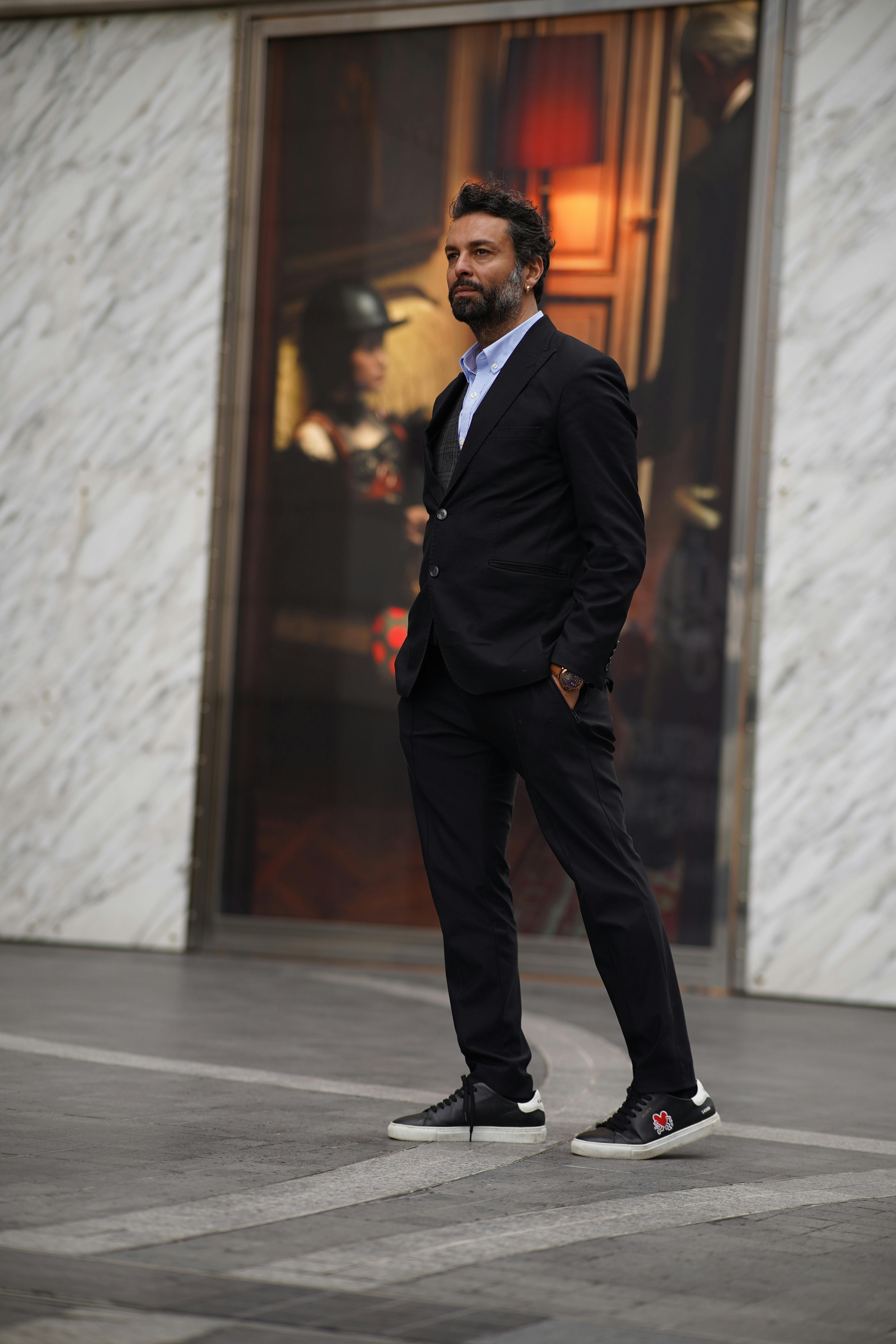 A fashionable bearded man in a black suit stands confidently on an urban street, exuding modern style.