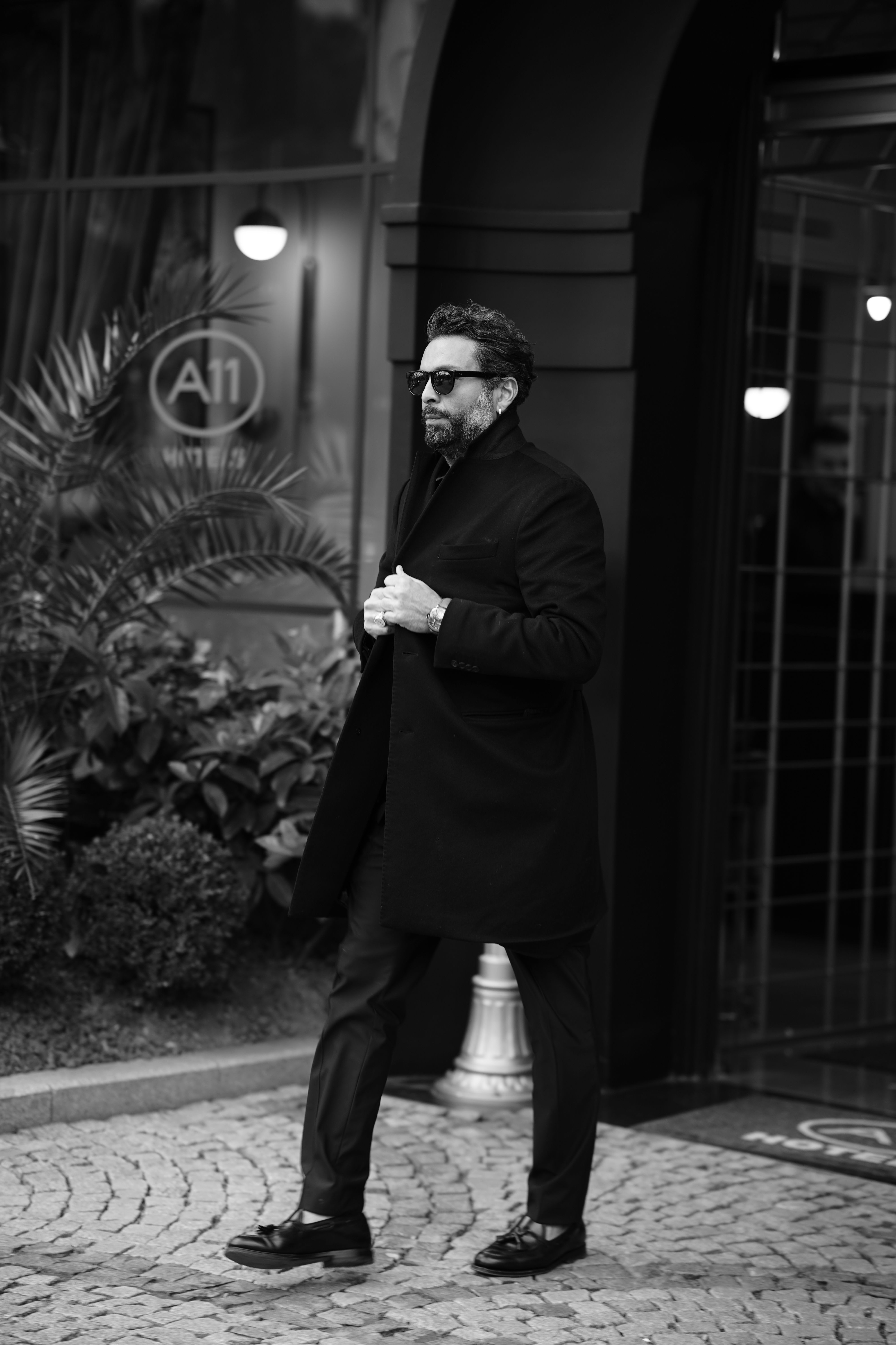 Man in a black coat and sunglasses walking outdoors in monochrome imagery.