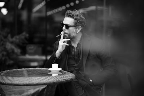 Grayscale Photo of a Man in Black Suit Wearing Sunglasses while Smoking Cigarette
