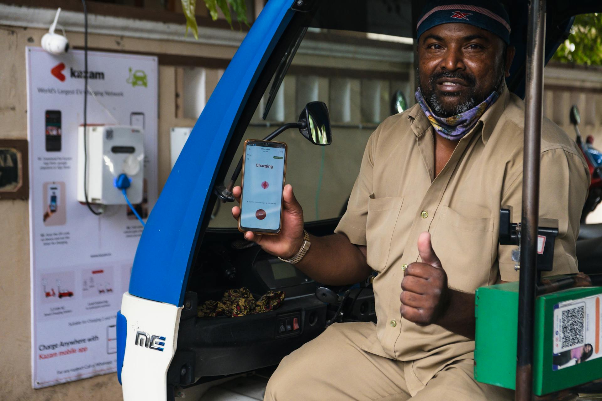 South Asian man using electric vehicle charging station in India, holding smartphone with app.