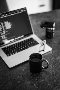 Black and white image of a laptop, coffee mug, glasses, and camera on a desk.