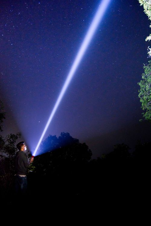 Free A man shines a bright flashlight into the starry night sky, creating a dramatic light beam. Stock Photo