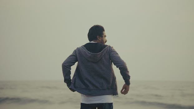 A man in a gray hoodie enjoys a serene beach sunset, facing the ocean.